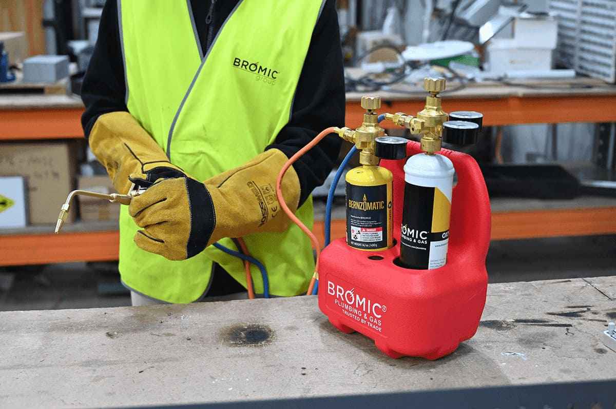 A person wearing a bright safety vest and gloves holds a blowtorch. Nearby, two gas cylinders labeled 