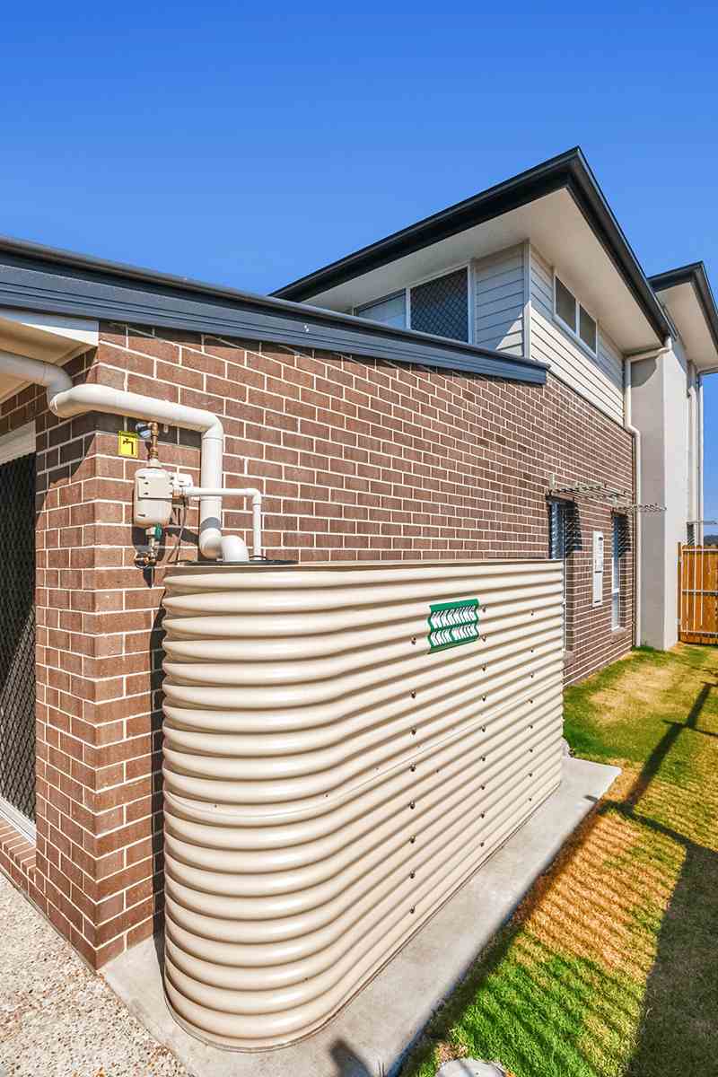 A beige, corrugated rainwater tank is installed against a brick house wall. Above, pipes are attached to the exterior. The context is a suburban yard on a sunny day.