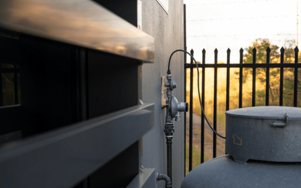 A close-up view of a gas regulator and piping system mounted on an exterior wall, with a hose connection leading to a gas storage tank. A black metal fence and a sunset-lit landscape are visible in the background